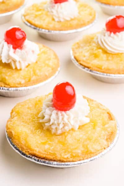A pineapple tart has whipped cream and a maraschino cherry on top. There are more of the tarts in the background.