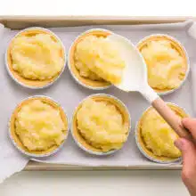 A hand holds a spatula, spooning pineapple filling into tarts in a baking pan.