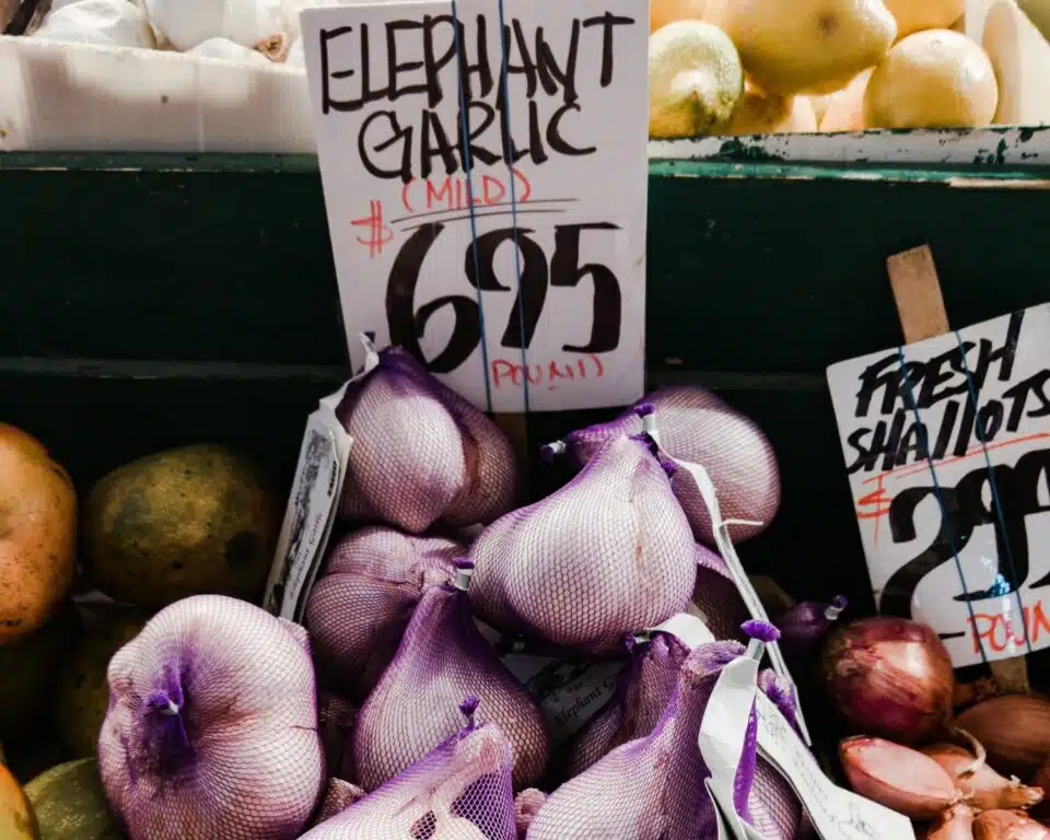 A farmer's market display has garlic wrapped in purple bags. The sign reads, Elephant Garlic, $6.95 per pound.