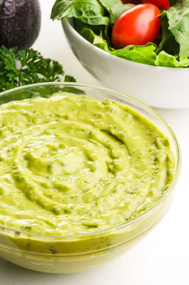 A bowl of avocado green goddess dressing sits in front of a salad and an avocado.
