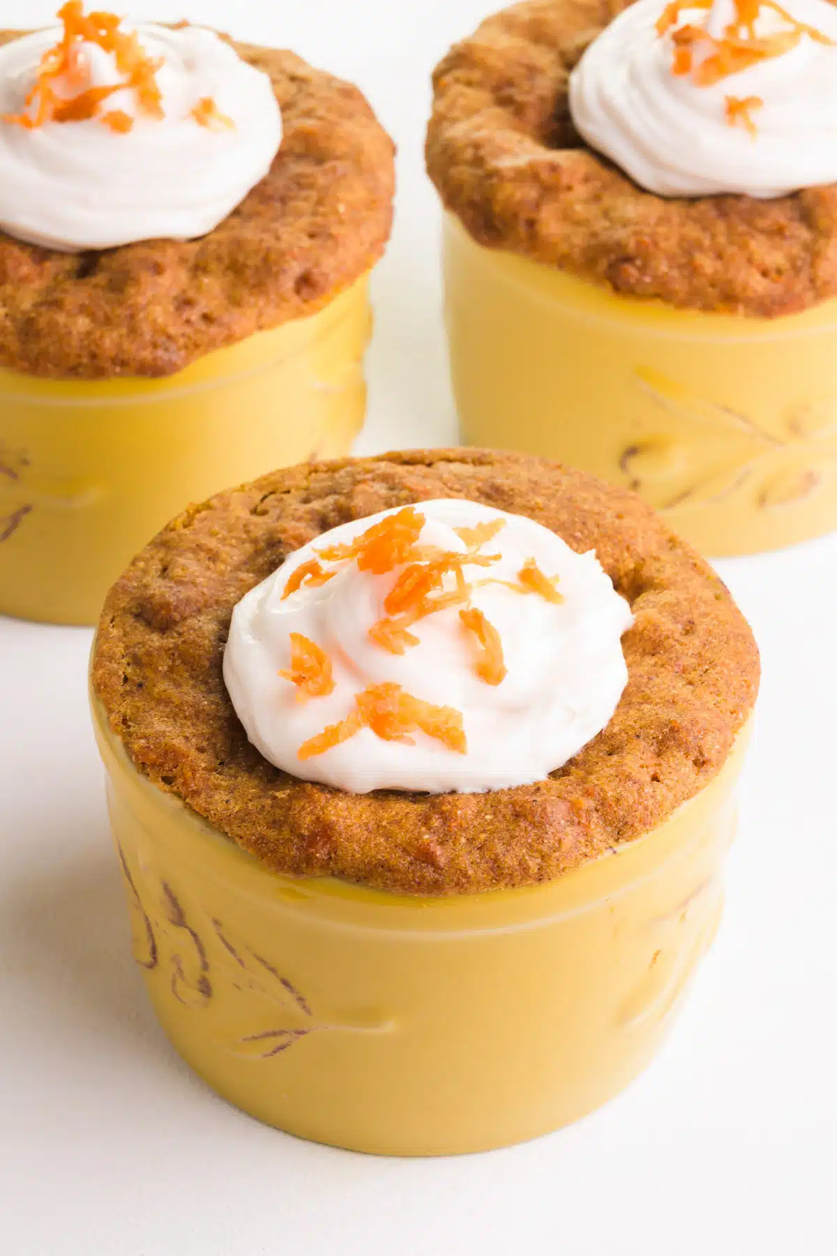 Three dishes of carrot mug cake have frosting and carrot shavings on top.
