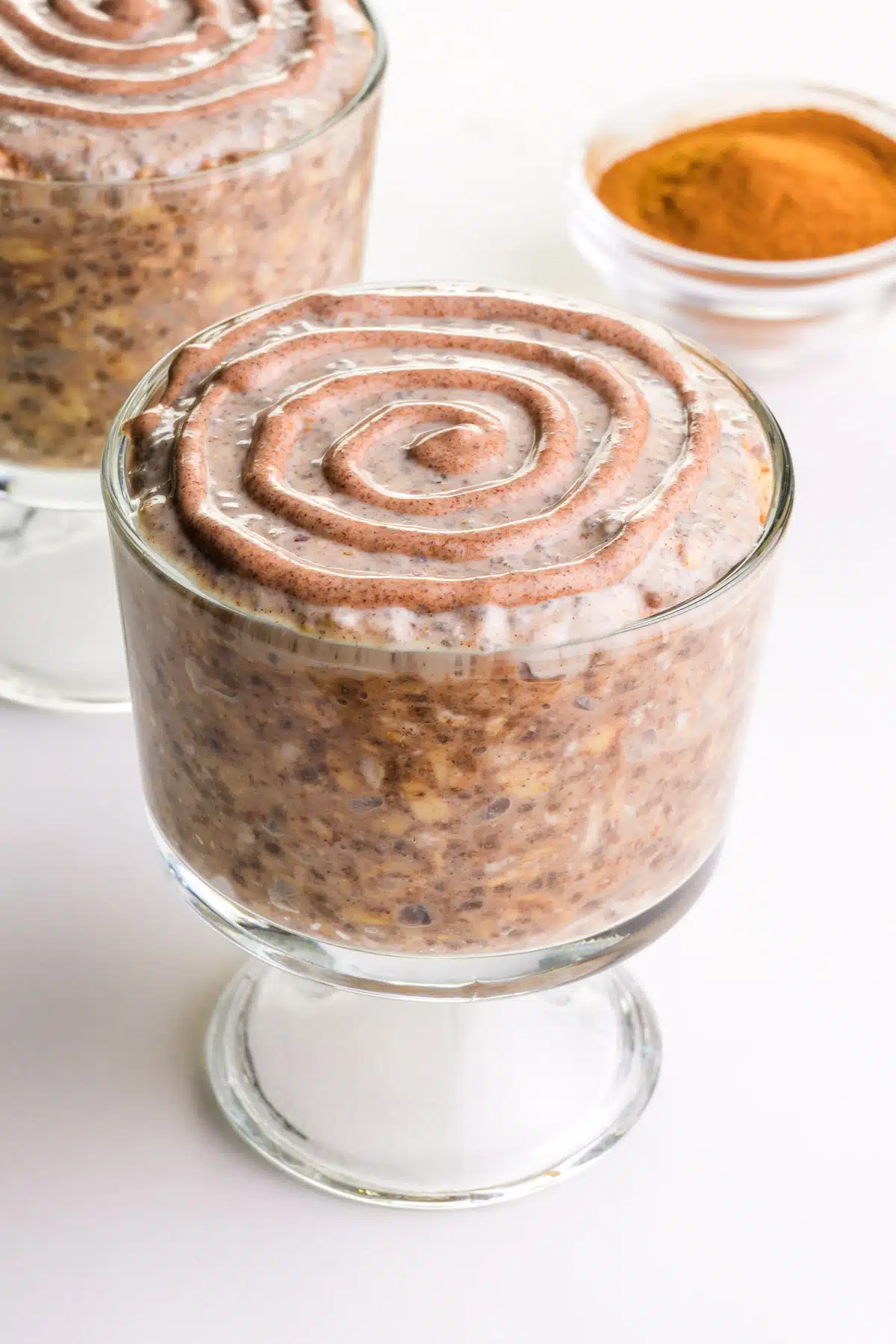 Two bowls of cinnamon roll overnight oats sits beside a bowl of ground cinnamon.