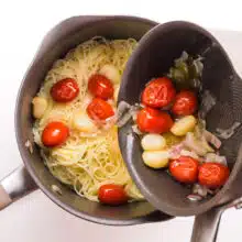 Roasted garlic, tomatoes, and onion, are being poured from one saucepan into another one with cooked pasta.