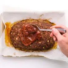 A hand holds a spoon, pouring tomato sauce over a baked meatloaf in a pan.