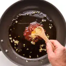 A hand holds a wooden measuring spoon, adding ingredients to a saucepan with garlic and sauce.