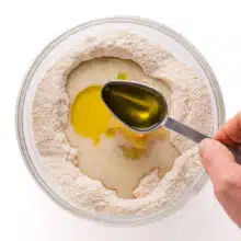 A hand pours a spoonful of olive oil into a bowl with other batter ingredients.