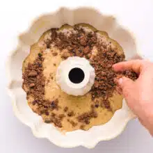 A hand distributes chocolate streusel over cake batter in a bundt pan.