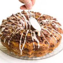 A hand holds a spoon drizzling vanilla icing over a coffee cake.