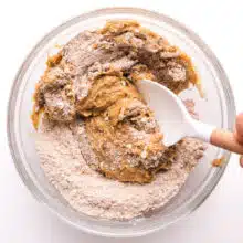 A hand holds a spatula stirring together flour ingredients with a wet mixture.