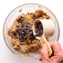 A hand holds a measuring cup of chocolate chips, pouring it into a bowl with cookie dough.