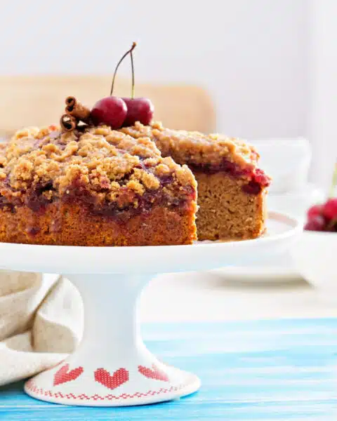 A coffee cake with cherries on top sits on top of a cake stand. It sits on a table with a blue table cloth.