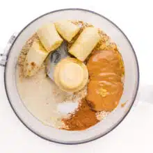 Looking down on a food processor bowl with ingredients such as sliced bananas, peanut butter, spices, and more.