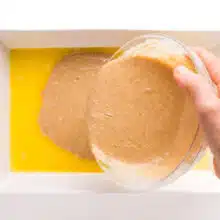 A hand holds a bowl pouring batter into a baking dish with melted butter.
